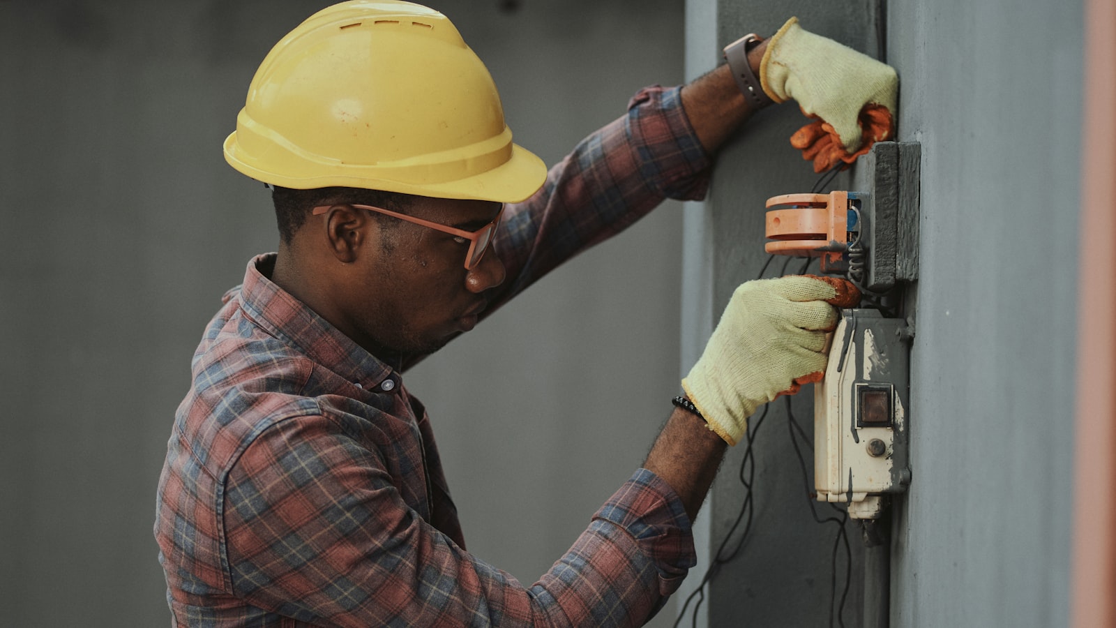 An electrician in a hard hat and gloves repairing an electrical panel on a building wall.